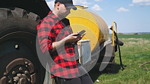 Young attractive farmer with phone standing in field, tractor working in green field in background. Smart farming using