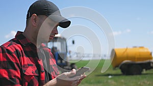 Young attractive farmer with phone standing in field, tractor working in green field in background. Smart farming using