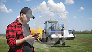 Young attractive farmer with phone standing in field, tractor working in green field in background. Smart farming using