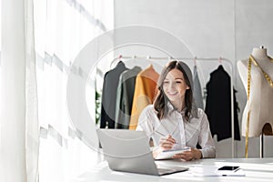 Young attractive female fashion designer sketching idea on paper at desk, working with a laptop at home