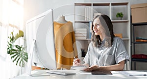 Young attractive female fashion designer sketching idea on paper at desk, working with a laptop at home