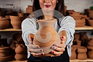 Young and attractive female potter working in a studio with a brown clay vase pot in the craft workshop holding the tool