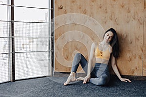 Young attractive fitness girl sitting on the floor near the window on the background of a wooden wall, resting on yoga classes