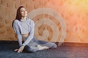 Young attractive fitness girl sitting on the floor near the window on the background of a wooden wall, resting on yoga classes