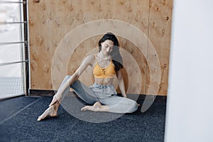 Young attractive fitness girl sitting on the floor near the window on the background of a wooden wall, resting on yoga classes