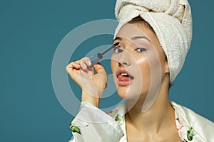 Young attractive funny woman putting eye mascara, studio shot over blue background, beauty treatment