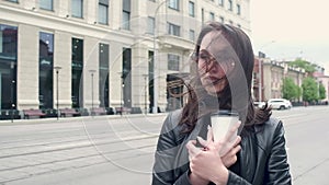 Young attractive girl in a black leather jacket and glasses stands in the cold wind with coffee in her hand. straightens hair. cit