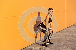 Young attractive girl in black sporwear practicing sport exercise on street, standing on squat near orange wall background, take