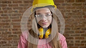 Young attractive girl builder is nodding her head and glances away, watching at camera, brick background