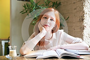 Young attractive girl female student with white skin and long red hair is reading books, studying, surrounded by books