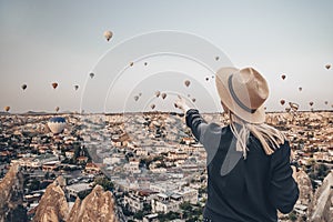 Young attractive girl in a hat stands on the mountain with flying air balloons on the background. Finger pointing girl