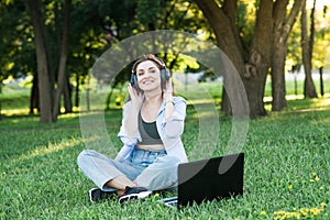 A young and attractive girl in headphones sitting on the grass on the lawn. Using laptop to listening to music with headphones.