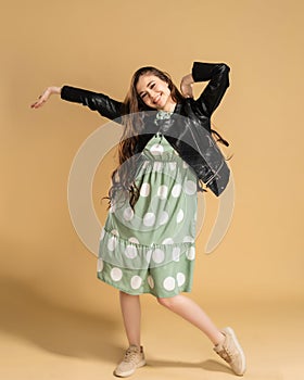 Young attractive girl with long curly hair in a dress with polka dots and a black leather jacket posing on an orange studio backgr