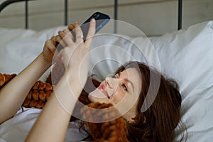 A young attractive girl is lying in bed with a smartphone, portrait, leafing through the news on the Internet and smiling