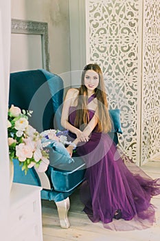 Young attractive girl in a magical charming light lush dress of purple color sits on a blue sofa and holds a stylish