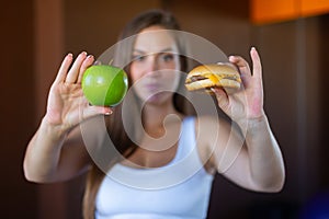 Young attractive girl is making choice between healthy and harmful food holding a green apple and a burger in her hands