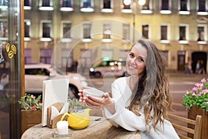 Young attractive girl sitting in the evening in a cafe with a Cup of tea to the backdrop of passing cars and city life. She looks