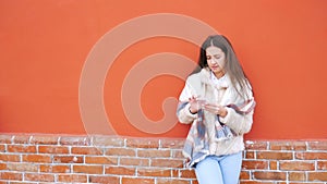 A young attractive girl stands at the wall of a building and pokes a finger into a mobile phone.