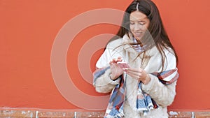 A young attractive girl stands at the wall of a building and pokes a finger into a mobile phone.