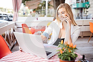 Young attractive girl talking on mobile phone and smiling while sitting alone in coffee shop during free time and working on table