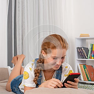 Young attractive girl teenager with red hair  lies on a sofa at home with a mobile phone.