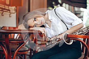 Young attractive girl in white shirt with a saxophone sitting on caffe shop - outdoor in street. young woman with sax looking