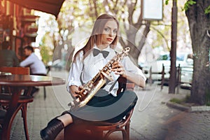 Young attractive girl in white shirt with a saxophone sitting near caffe shop - outdoor in sity. young woman with sax looking