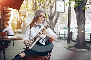 Young attractive girl in white shirt with a saxophone sitting near caffe shop - outdoor in sity. young woman with sax thinkin