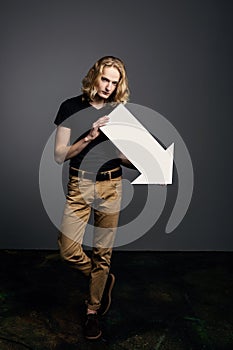 Young attractive guy with long blonde hair holds a large white arrow pointing down with a dejected face on a gray