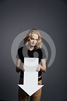 Young attractive guy with long blonde hair holds a large white arrow pointing down with a dejected face on a gray background