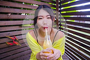 Young attractive and happy Asian Chinese girl having fun in garden outdoors drinking healthy orange juice with straw playful