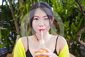 Young attractive and happy Asian Chinese girl having fun in garden outdoors drinking healthy orange juice with straw playful