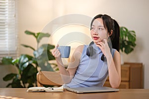 Young attractive happy Asian female student holding a cup of coffee sitting in the living room smiling and looking at