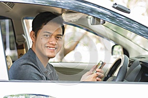 Young attractive happy asian male using mobile phone and looking at camera while driving his car in sunny day