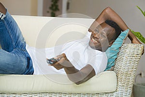 Young attractive and happy black African American man holding TV remote controller watching television movie enjoying relaxed