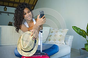 Young attractive and happy black hispanic woman at home with suitcase sitting on sofa couch leaving for holidays trip feeling exci