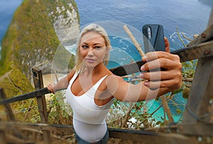 Young attractive and happy blond tourist woman taking selfie at beautiful sea cliff beach viewpoint enjoying exotic holidays