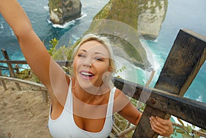 Young attractive and happy blond tourist woman taking selfie at beautiful sea cliff beach viewpoint enjoying exotic holidays