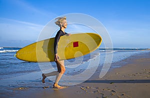 Young attractive and happy blonde surfer girl in beautiful beach carrying yellow surf board running out of the sea enjoying summer