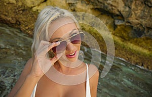 Young attractive happy and cheerful blond girl in bikini smiling playful holding sunglasses posing carefree at beautiful beach