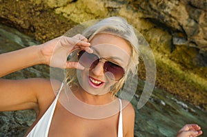 Young attractive happy and cheerful blond girl in bikini smiling playful holding sunglasses posing carefree at beautiful beach