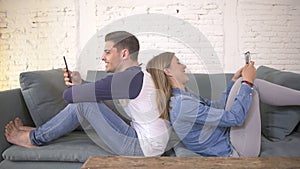 Young attractive and happy couple using internet app on mobile phone enjoying and laughing together sitting back to back at home c