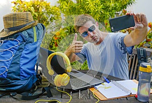 Young attractive and happy digital nomad man working outdoors with laptop computer cheerful taking selfie pic with mobile phone