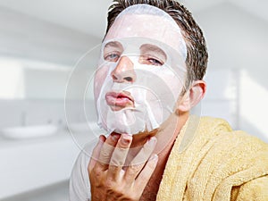 Young attractive and happy gay man in bathroom using beauty paper facial mask touching his face looking to the mirror smiling in