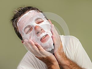 Young attractive and happy gay man in bathroom using beauty paper facial mask touching his face looking to the mirror smiling in