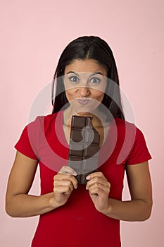 Young attractive and happy hispanic woman in red top smiling excited eating chocolate bar on pink background