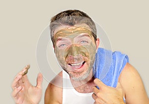 Young attractive and happy man with bathroom towel laughing of himself in the mirror with green cream on his face applying facial