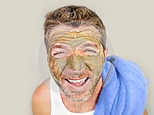 Young attractive and happy man with bathroom towel laughing of himself in the mirror with green cream on his face applying facial