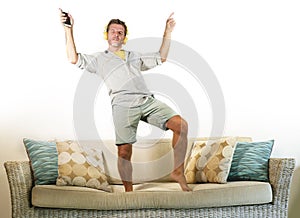 Young attractive and happy man with headphones and mobile phone listening to music jumped on sofa couch dancing and singing crazy