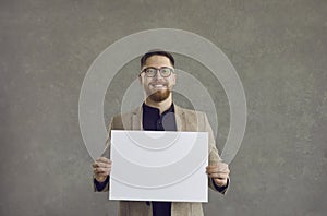 Young attractive happy man holding a blank white sheet while standing on a gray background.